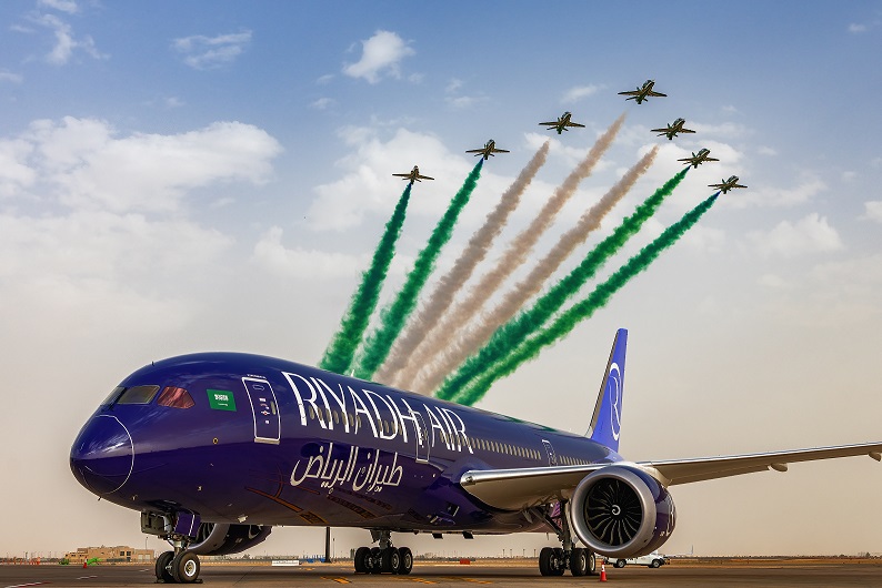 A Riyadh Air plane in the foreground, with an aeronautical display in the background.