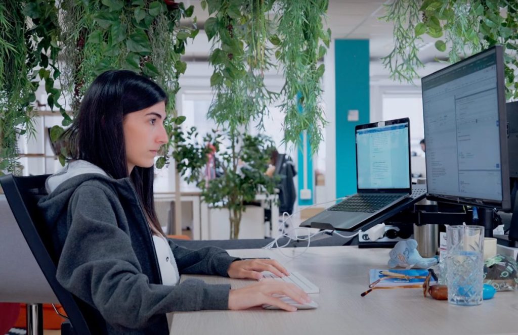 A woman working at a desk in an office looking at two computer monitors.