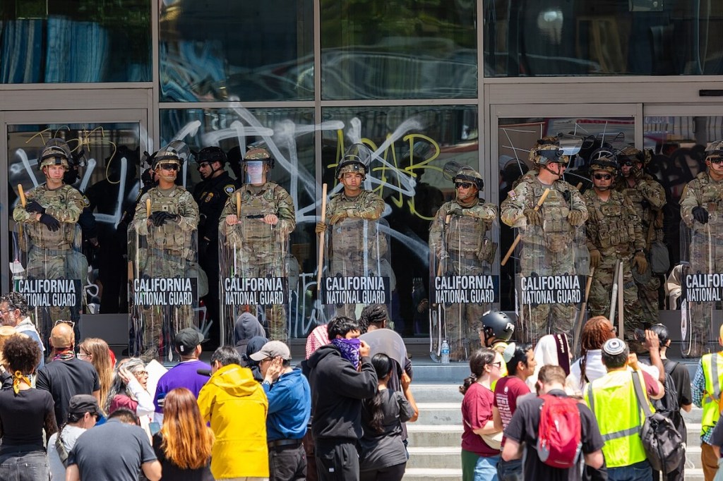 California National Guardsmen with protective shields standing on top of steps with pro-immigrant protesters gathered below.