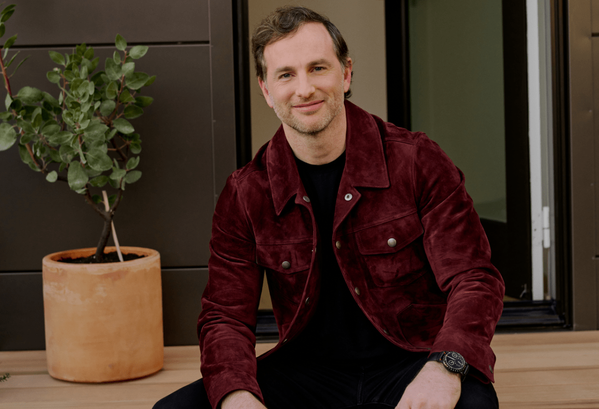 A man seated with a plant in the background.