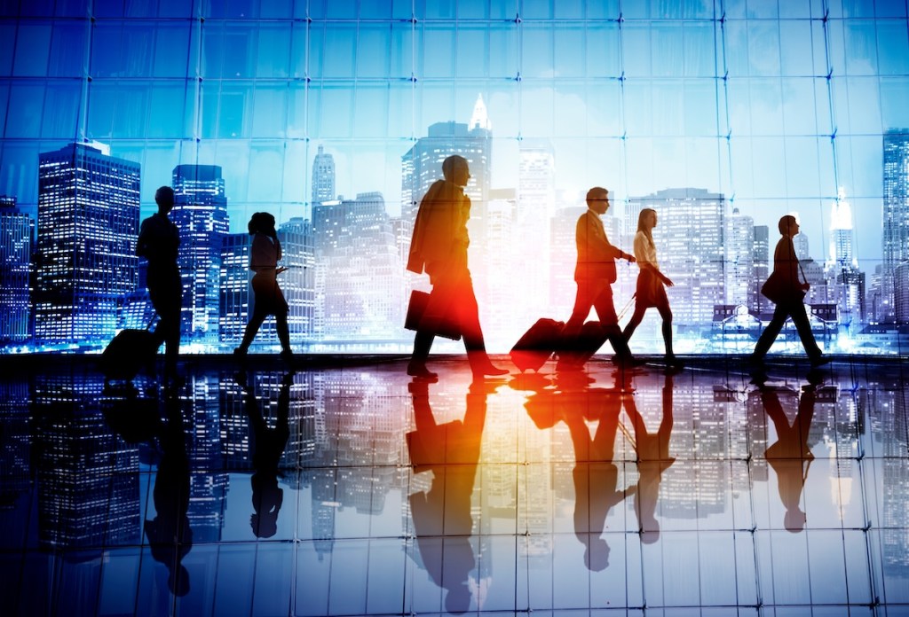 Business people walking in a large hall that could be an airport or possibly another transportation facility. There are buildings in the background, which is a bit hazy.