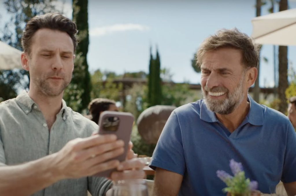 Two men sitting outdoors at a table with one checking a hotel price on his phone.