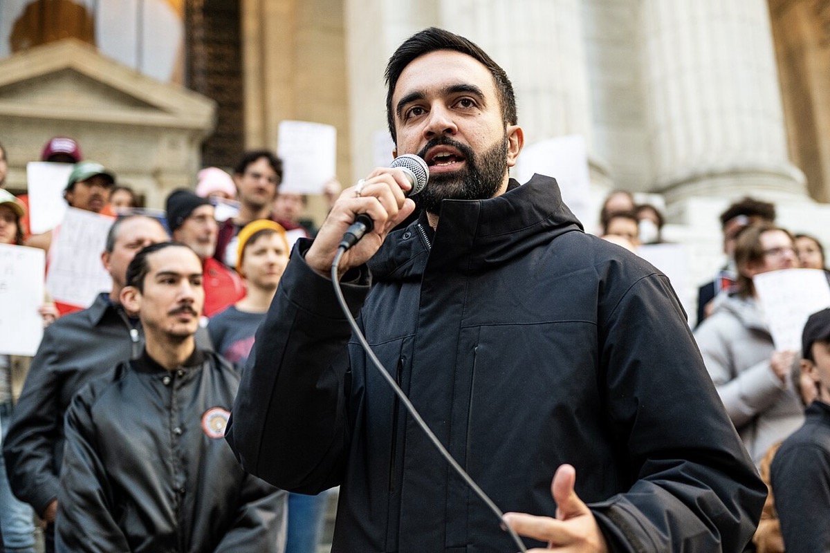 A man speaking on a microphone with people lined up behind him.