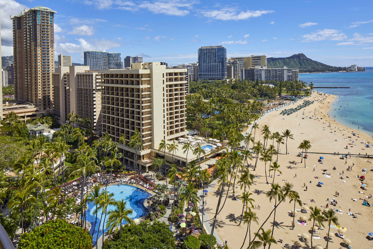 Image of a beach hotel in Waikiki Beach in Hawaii on a sunny day. View of a pool and happy guests shown from an aerial view so you can't tell what hotel it is.
