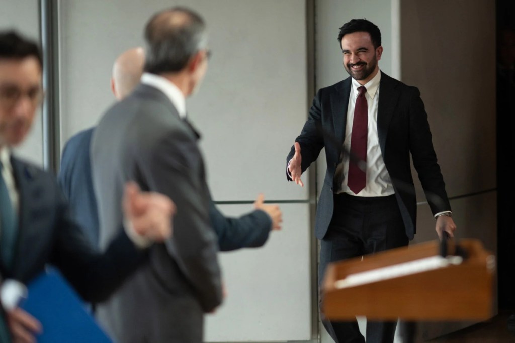 Image of Mayor Zohran Mamdani holding out his hand and shaking other officials' hands at a press conference.