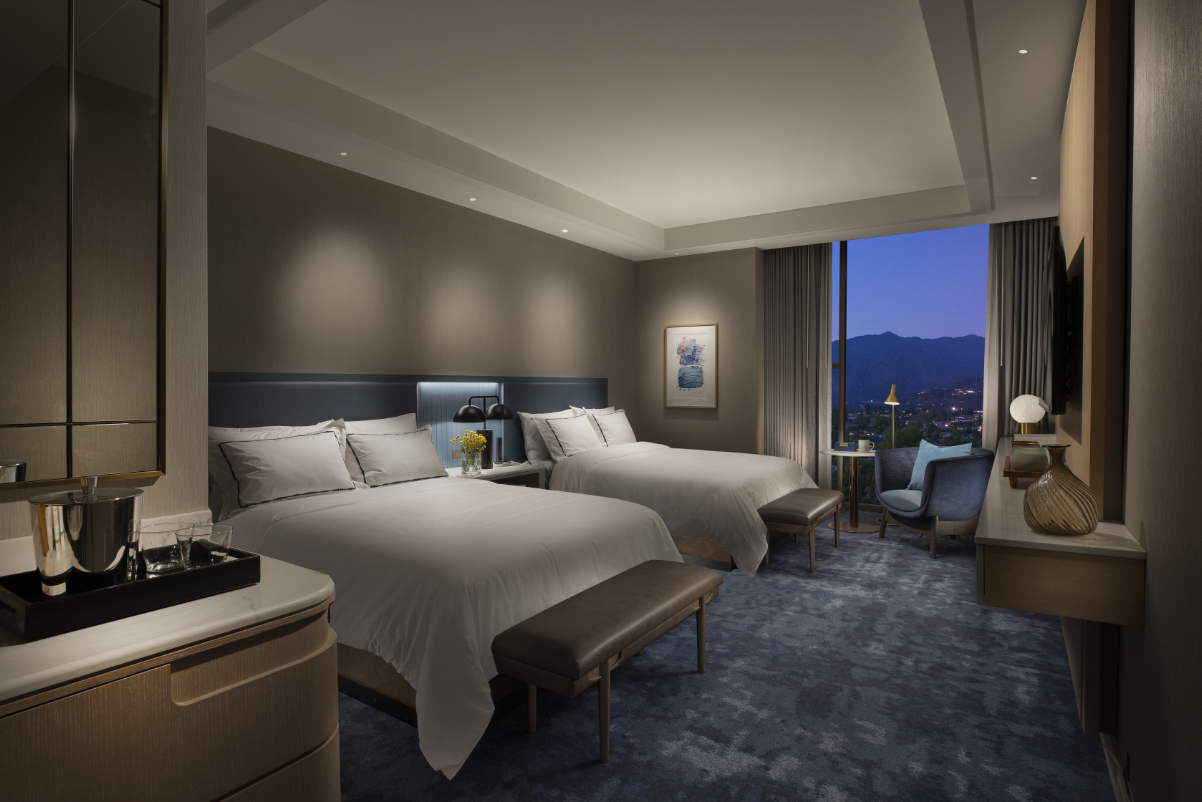 A duskily lit interior view of a double bed guest room in a newly renovated Hilton in Los Angeles with a view in the evening outside the window.