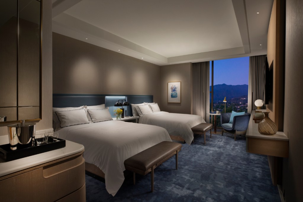 A duskily lit interior view of a double bed guest room in a newly renovated Hilton in Los Angeles with a view in the evening outside the window.