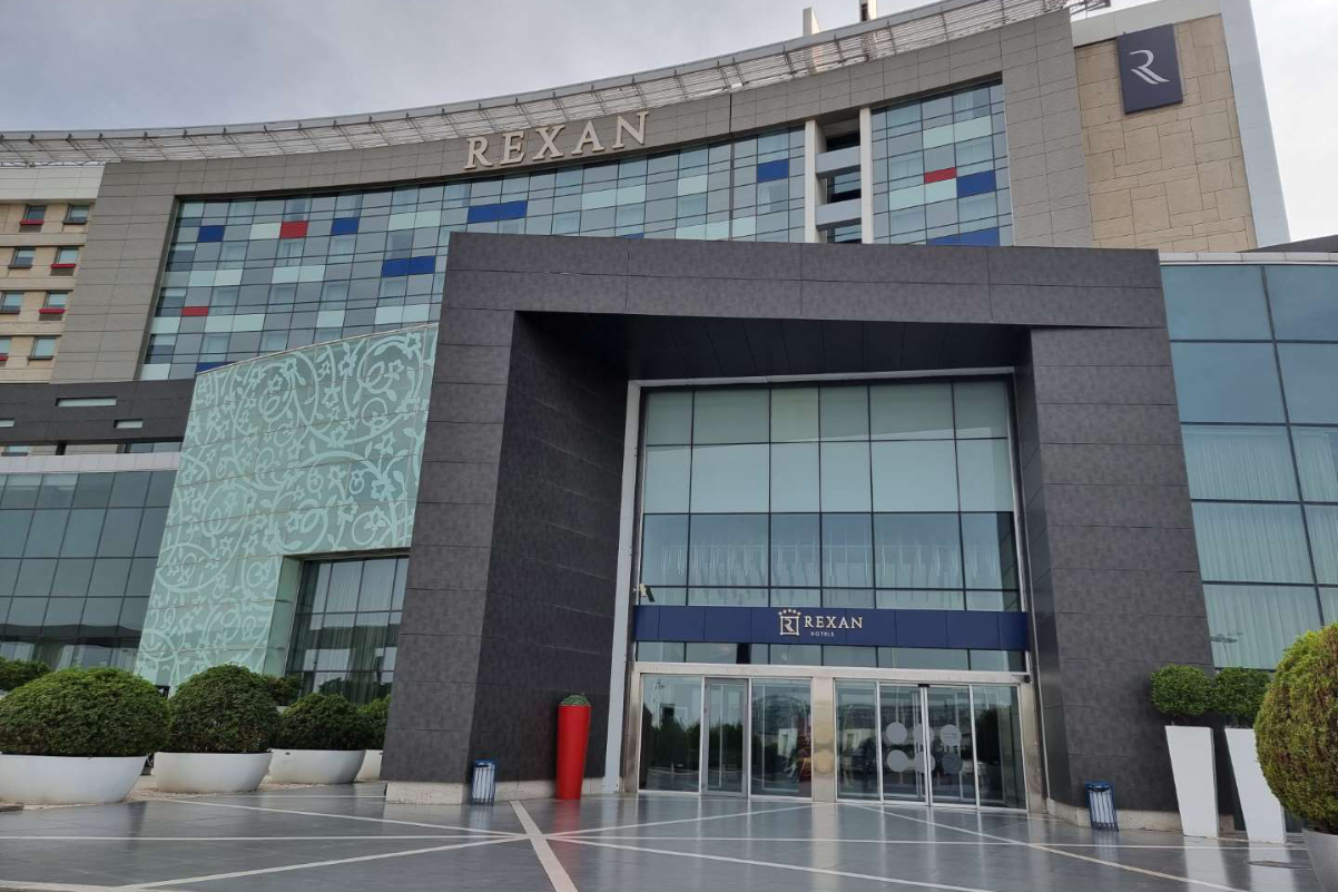 an exterior image of the Rexan Tehran Airport Hotel with distinctive Arabic artistry on the glass and concrete building by the airport on a sunny day