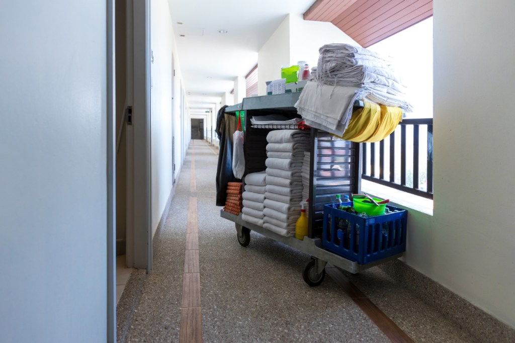 Image of a cart with towels and other hospitality items for cleaning and refreshing a guest room at a hotel, sitting alone in a corridor that's brightly lit.