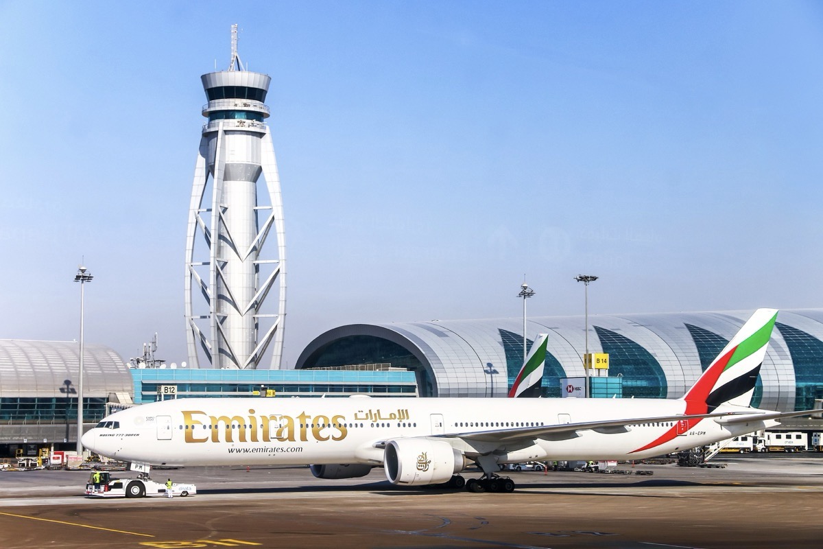 A plane taxiing on an airport runway with a tower and a terminal in the background.