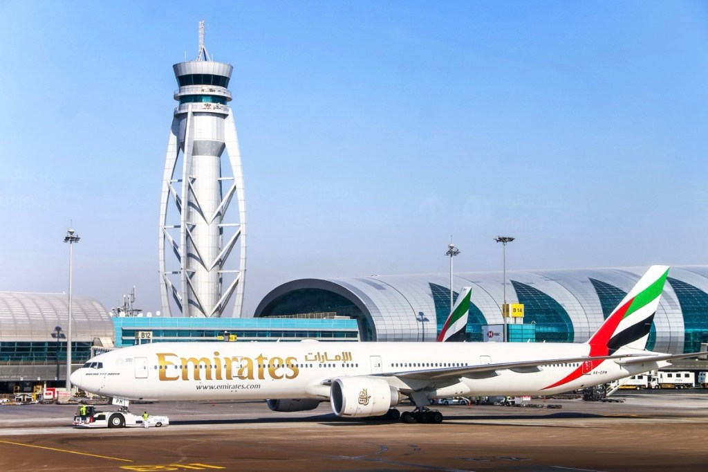 A plane taxiing on an airport runway with a tower and a terminal in the background.