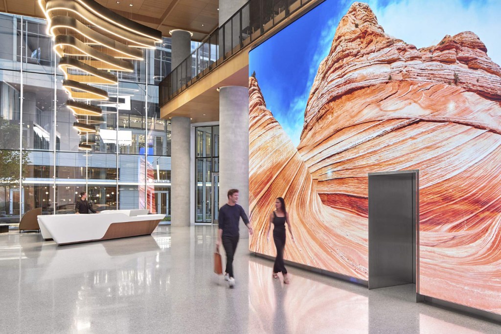 Image of a lobby of an office building, namely the Marriott headquarters, with a giant LED screen showing a dramatic visualization of a mountain rock outcrop, with some employees walking past.