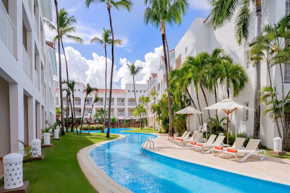 View of an all-inclusive resort showing a lazy river running through, with palm trees and deck chairs, and a sunny sky.