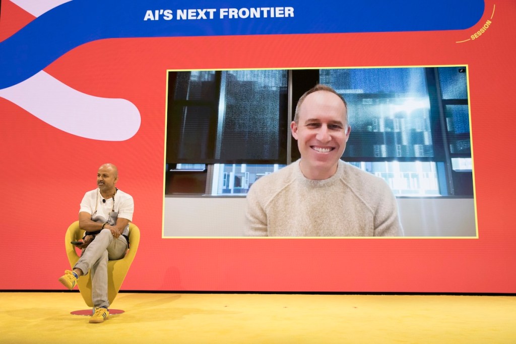 a man sits on a stage while interviewing another man on a screen behind him.
