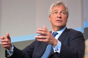 A man in a suit with a blue tie seated and holding his hands out in front of him, gesturing.