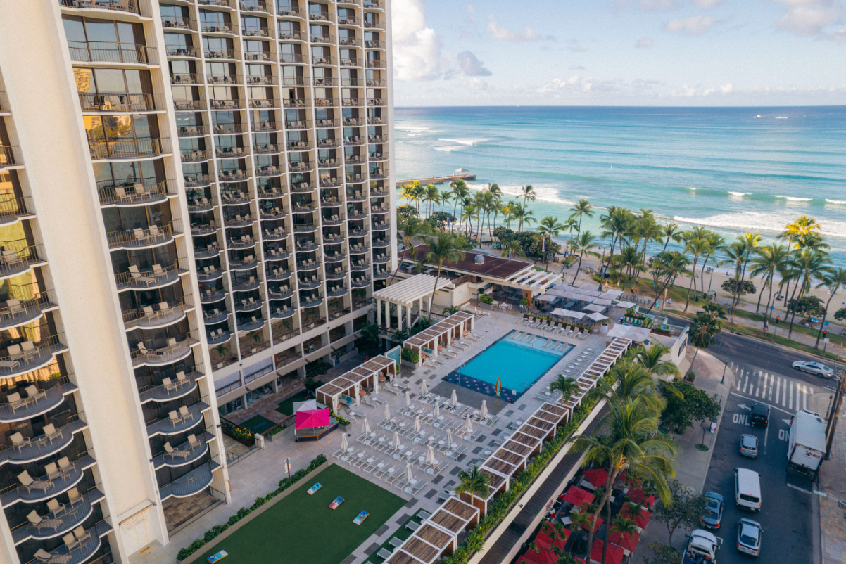 Image of a married hotel overlooking Waikiki Beach, seen from an aerial view of the hotel's pool, then the beautiful palm trees and ocean