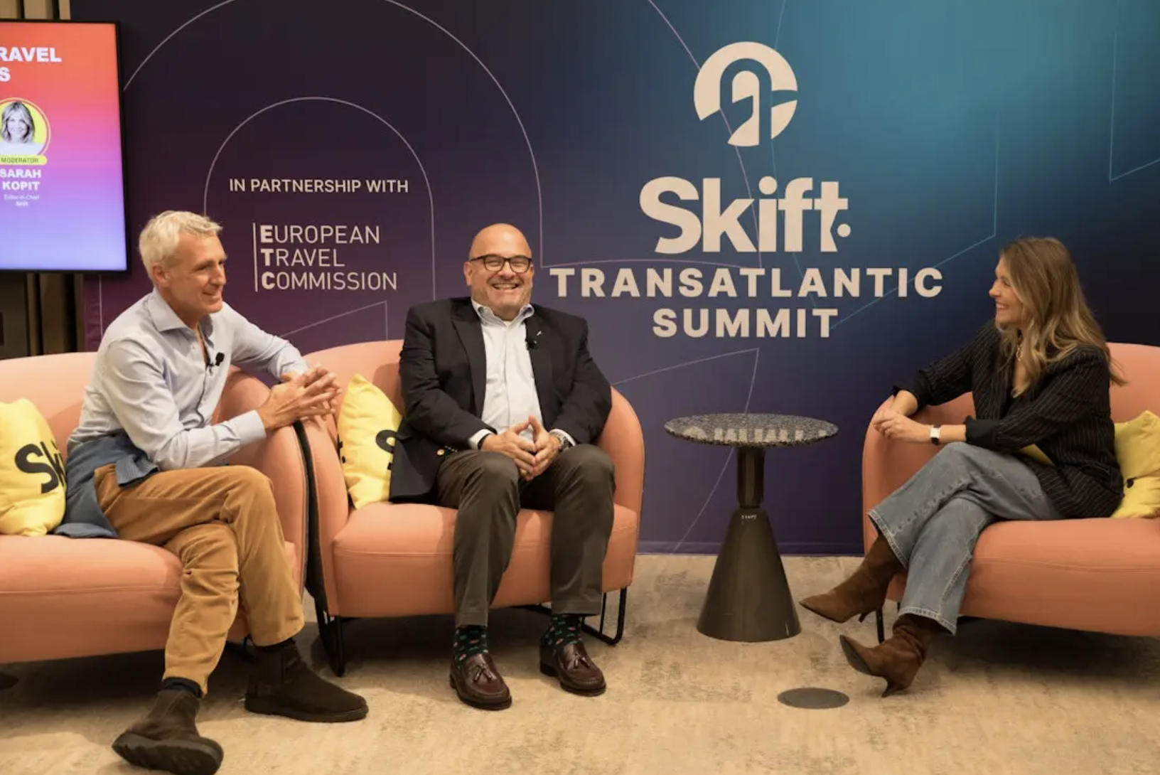 Three people sit and converse on stage at the Skift Transatlantic Summit, with event branding and sponsor logos visible in the background.