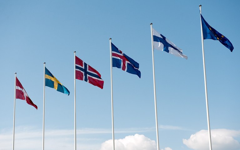 A set of European country flags flying on poles to a blue sky in the background.